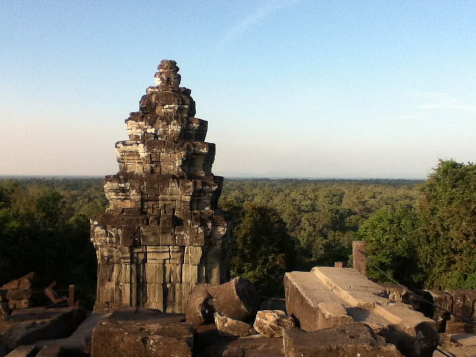 A weathered, stone temple structure rises from the ancient ruins, its worn and uneven layers revealing the passage of time. The temple, set against a vast, green expanse of forest that stretches to the horizon, contrasts with the blue sky above. The soft, golden light of the early morning or late afternoon bathes the scene, highlighting the rich textures of the stone and the lush greenery in the distance. The serene atmosphere is further enhanced by the quiet solitude of the location, with a single person seated quietly near the structure, blending into the timeless setting.