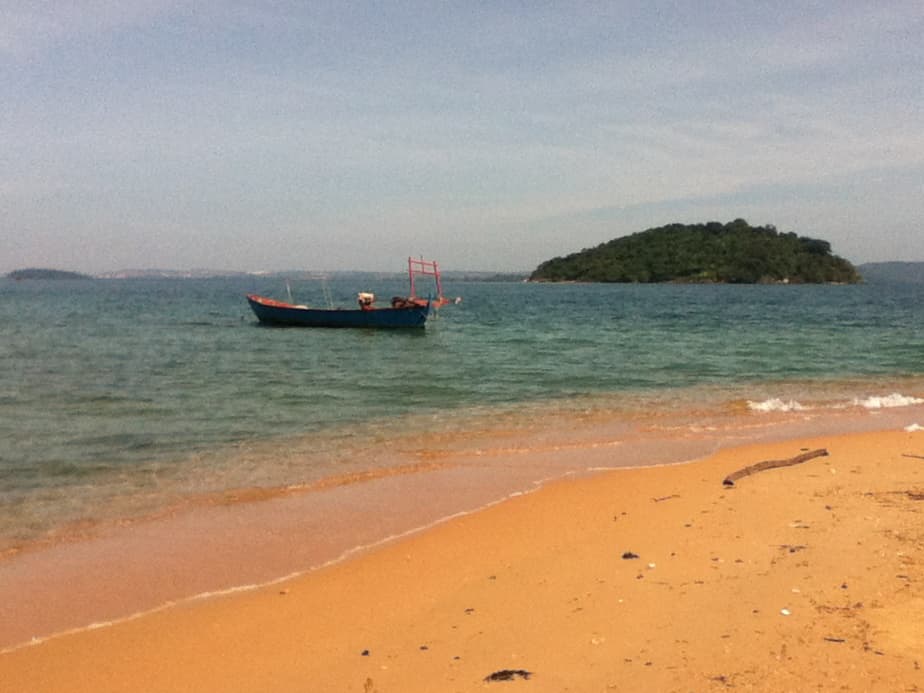 A serene beach scene with a small wooden boat anchored in shallow, clear waters near the shore. The sandy beach is warm and golden, with gentle waves washing up onto it. In the background, there is a small, lush, tree-covered island surrounded by the calm sea. The sky is a light blue, adding to the tranquil atmosphere of the scene.