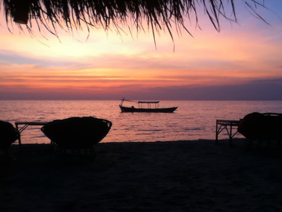 A serene beach scene at sunset with a small, traditional boat floating on calm waters. The sky is painted with soft pinks, oranges, and purples, casting a warm glow over the horizon. Silhouettes of wicker chairs and tables are visible in the foreground under the shadow of a thatched roof, adding to the peaceful and relaxing atmosphere. The boat, gently anchored, sits against the vast backdrop of the sunset-drenched sea.