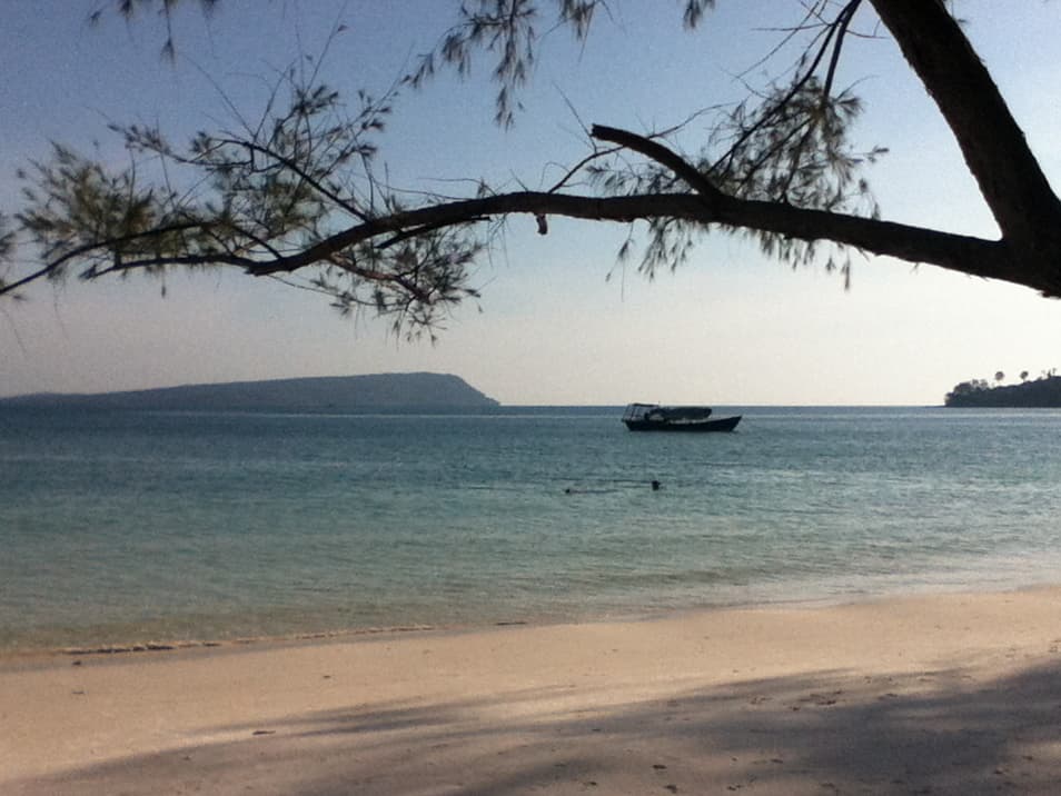 A peaceful beach scene with a tree branch framing the top of the image, casting shadows on the soft, sandy shore. In the distance, a boat is anchored on the calm, blue water. An island is visible across the horizon under a clear sky, creating a tranquil and serene atmosphere.