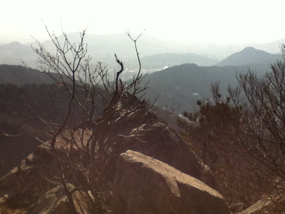This image shows a mountainous landscape with rocky terrain in the foreground and a misty, distant view of hills or mountains in the background. Sparse trees with bare branches frame the rocky outcrop, giving it a sense of natural isolation and calm. The atmosphere seems hazy, possibly from fog or mist, with city buildings faintly visible in the distance, adding a contrast between nature and urban life.