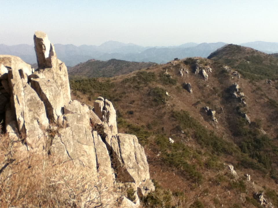 This image captures a rugged mountainous landscape with prominent rock formations in the foreground. The jagged cliffs and large boulders dominate the left side of the frame, while rolling hills with sparse vegetation extend into the background. The distant mountains are layered with a faint haze, giving the scene a sense of vastness and depth. The dry, brownish tones of the vegetation suggest that the photo may have been taken during a cooler or drier season. The contrast between the rocky cliffs and the softer, distant hills adds a dynamic quality to the landscape.