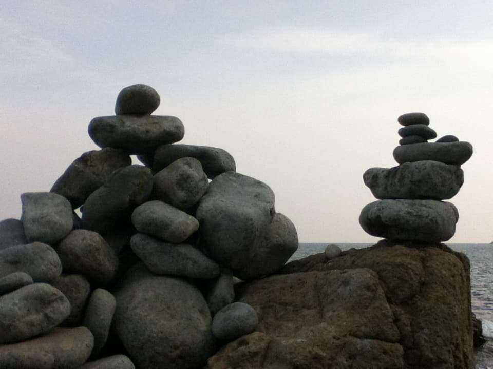 This image shows two stacks of balanced stones, also known as cairns, placed on a rocky outcrop by the sea. The sky is overcast, with soft light casting minimal shadows, and the calm ocean stretches into the horizon in the background. The stones are rounded and weathered, likely shaped by natural forces such as water and wind. The cairns create a peaceful, meditative atmosphere, symbolizing balance and harmony with nature. The juxtaposition of the solid stones and the expansive sea enhances the sense of serenity in the image.