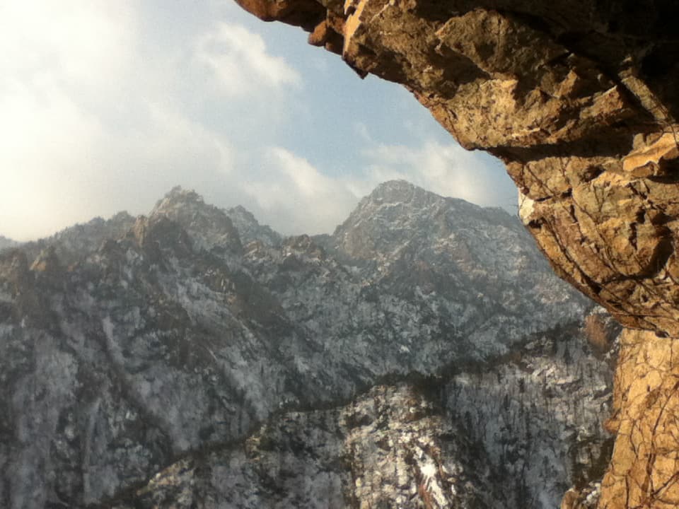 This image presents a breathtaking mountain landscape, with snow-dusted rocky peaks towering under a partly cloudy sky. The rocky overhang in the foreground adds a sense of depth and framing to the scene, with the rough texture of the cliff contrasting the smoother, snow-covered slopes of the mountains in the background. The interplay of light and shadow on both the rock formations and the mountains enhances the dramatic nature of the terrain. The image evokes a sense of awe and solitude, capturing the rugged beauty of a remote and elevated natural setting.