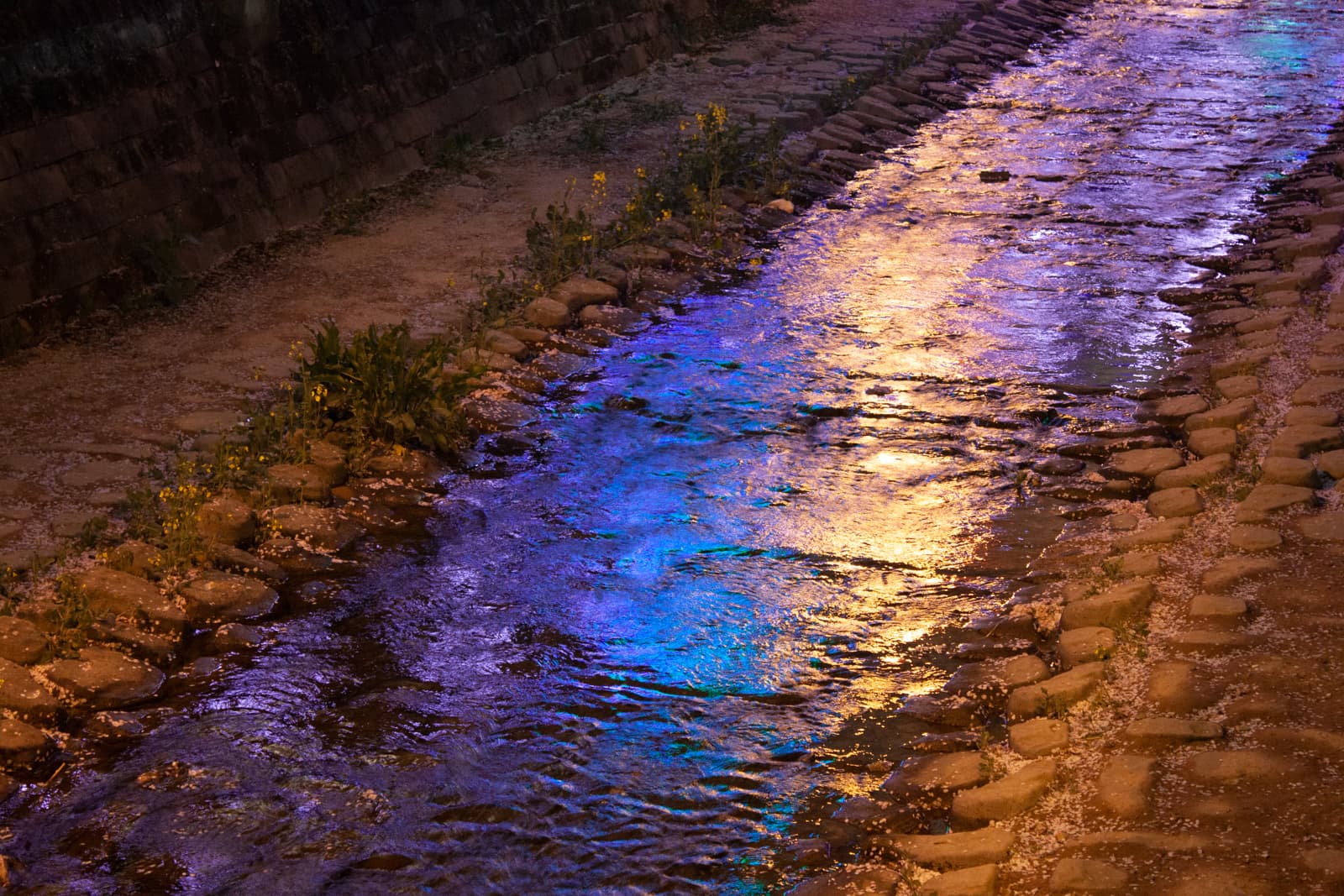 This image shows a narrow stream or waterway with cobblestones along its edges, illuminated by vibrant colors reflecting off the water's surface. The reflections, likely from nearby artificial lights, cast shades of blue, purple, and golden yellow, creating a striking and almost surreal effect. The gentle flow of the water is visible in the ripples that capture the shifting colors. Small patches of greenery grow along the cobbled edges, adding a natural touch to the otherwise structured setting. The scene exudes a calm, almost dreamlike atmosphere, with the play of light on water making it feel magical and serene.