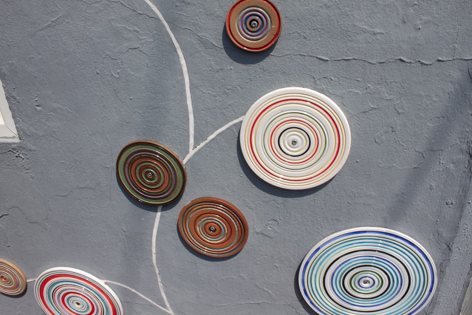 This image features a decorative installation of circular ceramic plates or discs mounted on a gray textured wall. Each disc has concentric rings in various colors, such as red, green, blue, and brown, creating a visually striking pattern. The design on the wall resembles an abstract tree, with white lines connecting the discs, mimicking branches or stems. The vibrant colors and geometric shapes stand out against the muted gray background, giving the installation a modern, artistic feel. The overall effect is playful and creative, adding an element of visual interest to the otherwise plain wall.
