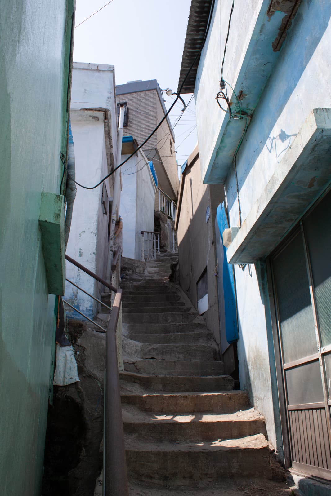 This image shows a narrow, steep staircase winding between closely packed buildings in what appears to be an urban residential area. The buildings on either side are worn, with faded paint and exposed concrete, creating a gritty and authentic atmosphere. Some of the walls are painted in pastel hues like light blue and green, while utility wires crisscross overhead, further emphasizing the lived-in, informal character of the space. The staircase leads upwards, drawing the eye towards the upper levels of the buildings, which seem to rise haphazardly as if stacked upon one another. The overall scene has a sense of quiet solitude and the charm of an older, densely populated neighborhood.