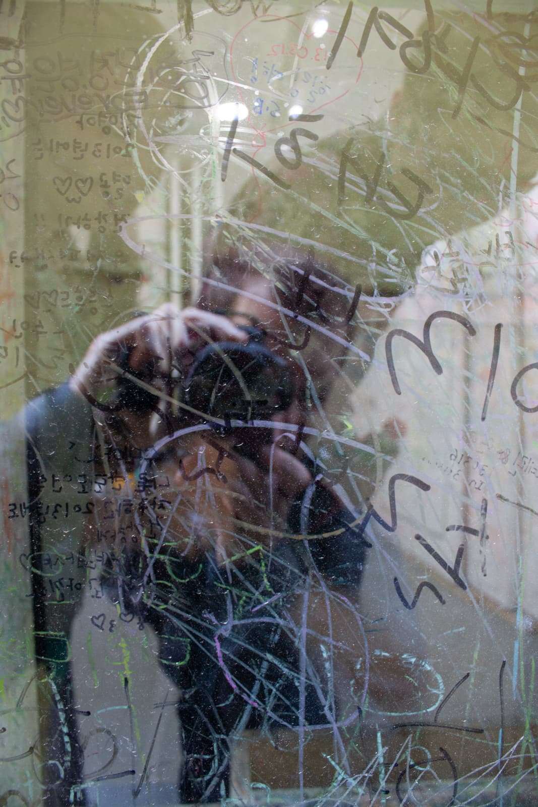 This image features a reflection of the photographer in a scratched and graffiti-covered glass surface. The glass is marked with various scribbles, drawings, and writings, creating an abstract and chaotic pattern. The layered effect of the reflections and the markings gives the image a sense of depth and mystery, as the viewer can partially make out the photographer’s form behind the glass. The interaction between the viewer and the artwork through the reflection suggests themes of identity, perception, and interaction with urban spaces, where public markings become part of the visual landscape. The overall effect is a mix of personal and impersonal, as the reflective surface both reveals and obscures at the same time.