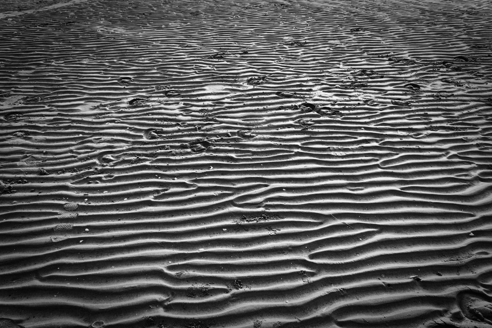 This black-and-white image depicts an expansive view of rippled sand, likely on a beach or desert. The smooth, repetitive patterns of the sand create a hypnotic texture, with each ripple catching the light and casting shadows that emphasize the undulating surface. Footprints and small marks scattered across the sand add a human element, subtly breaking the uniformity of the natural ridges. The interplay of light and shadow gives the scene depth and movement, making the simple sand ripples feel almost sculptural. The monochrome effect enhances the texture and form, transforming the ordinary into an abstract visual composition.