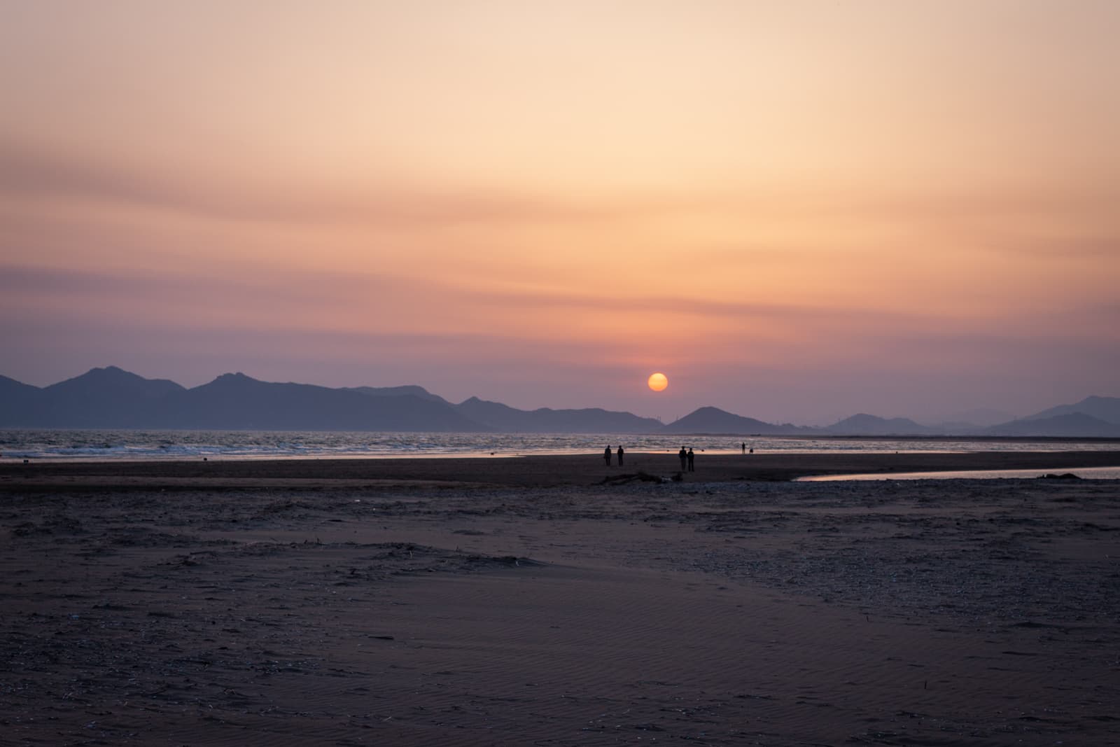 This image captures a serene sunset over a beach, with the sun hanging low in the sky as it casts a warm orange and purple glow across the horizon. The distant mountains are silhouetted against the soft gradient of colors in the sky, adding depth to the scene. The calm sea reflects the fading sunlight, creating a tranquil atmosphere. In the foreground, a few people can be seen walking along the beach, their small figures emphasizing the vastness of the landscape. The gentle waves and the expansive sand add to the peacefulness of the scene, evoking a sense of quiet reflection and beauty in nature.
