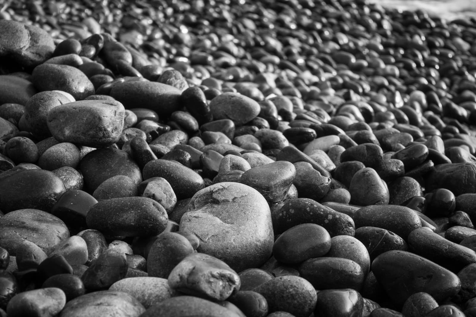 A close-up black-and-white shot of smooth, rounded pebbles covering a beach. The variety of stone sizes and the interplay of light and shadow across their surfaces emphasize the natural texture and form of the rocks.