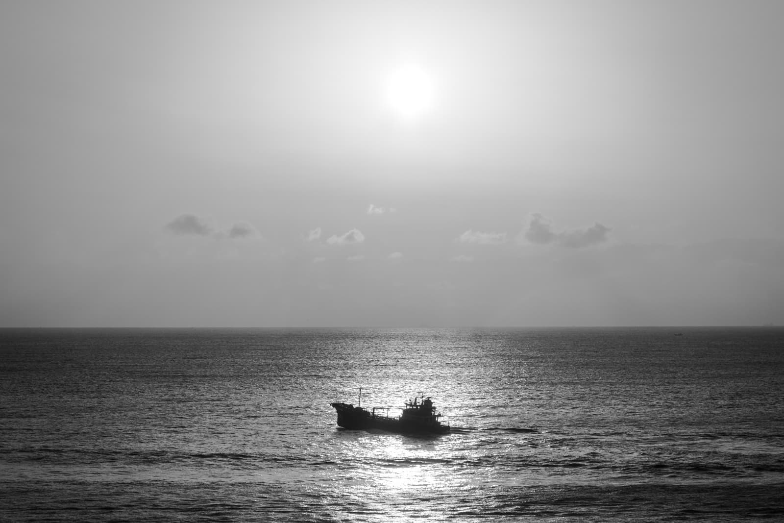 A black-and-white photograph of a lone boat on the ocean, with the sun shining high in the sky and reflecting off the water’s surface. The horizon is vast and uninterrupted, giving the image a sense of calm and solitude.