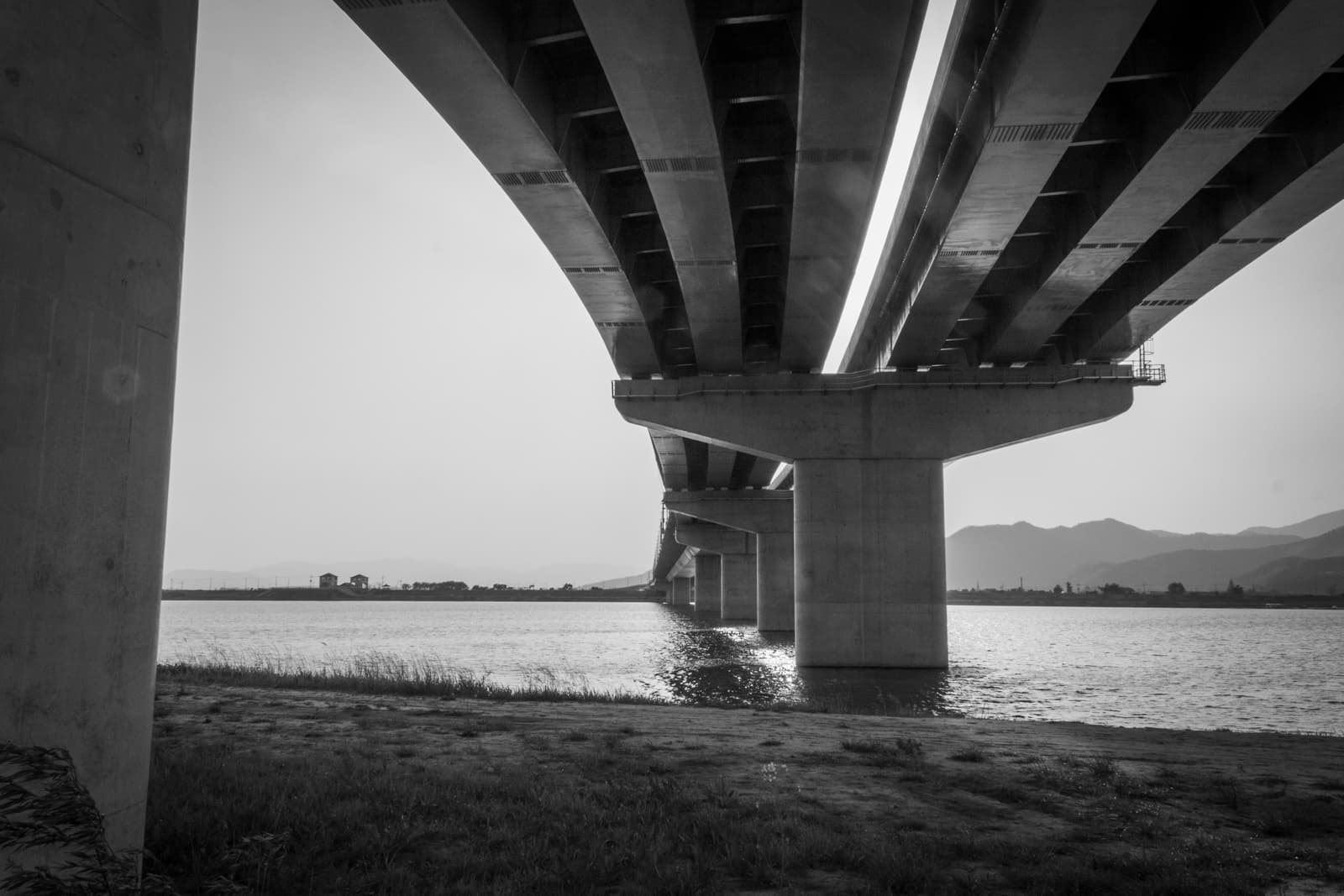 A black-and-white photograph of a large bridge, seen from below, with its concrete supports and the underside of the road stretching across a body of water. The perspective emphasizes the vastness and structural precision of the bridge, as it disappears into the distance.