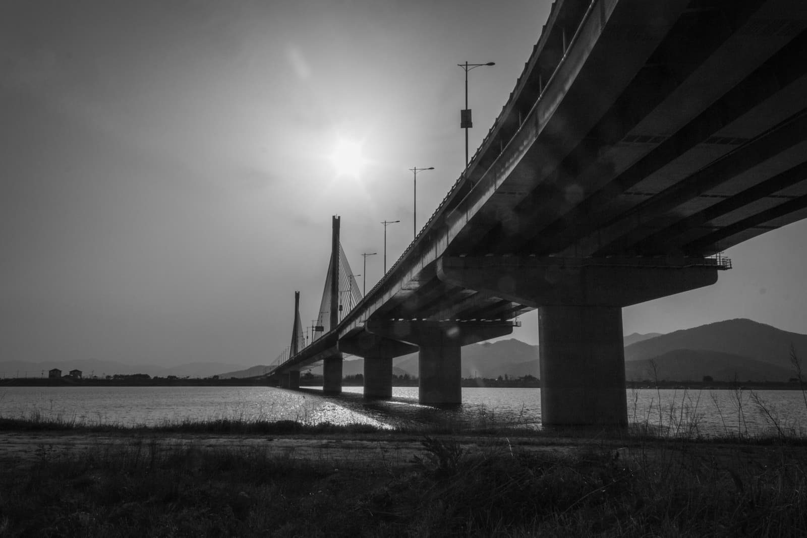 A black-and-white shot of a long bridge extending across the water, captured in silhouette against the sun. The sun is low in the sky, casting light over the bridge and creating a reflection on the water, giving the image a dramatic and peaceful tone.