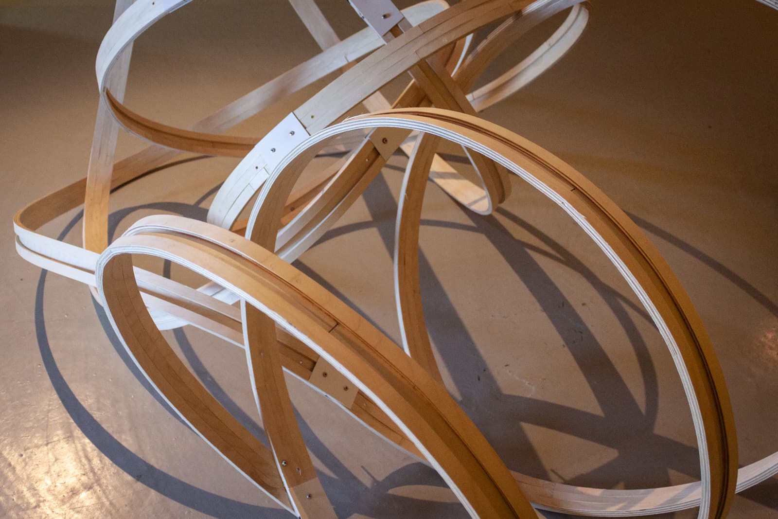 An abstract wooden sculpture of interlocking hoops placed in a gallery, casting shadows on the floor.