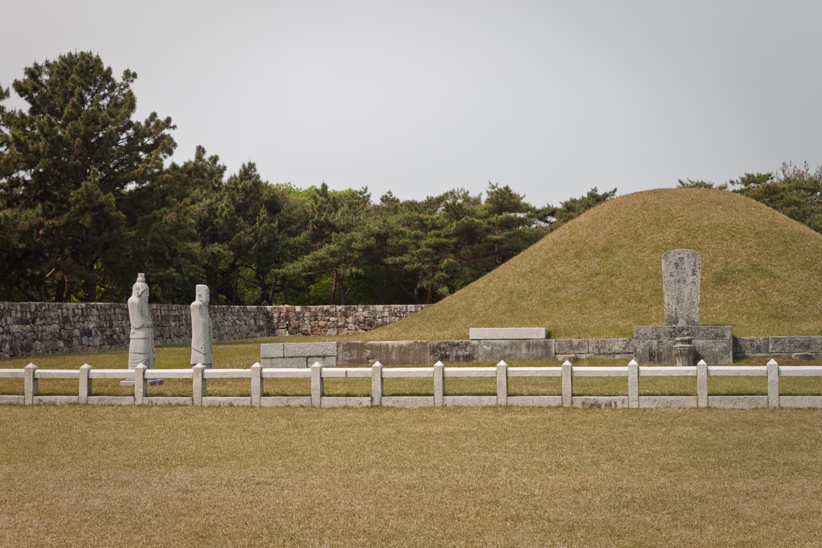 An ancient Korean royal tomb surrounded by well-maintained grounds. The burial mound is accompanied by statues of stone guards and a surrounding stone wall, indicative of traditional Korean burial sites of noble or royal figures.