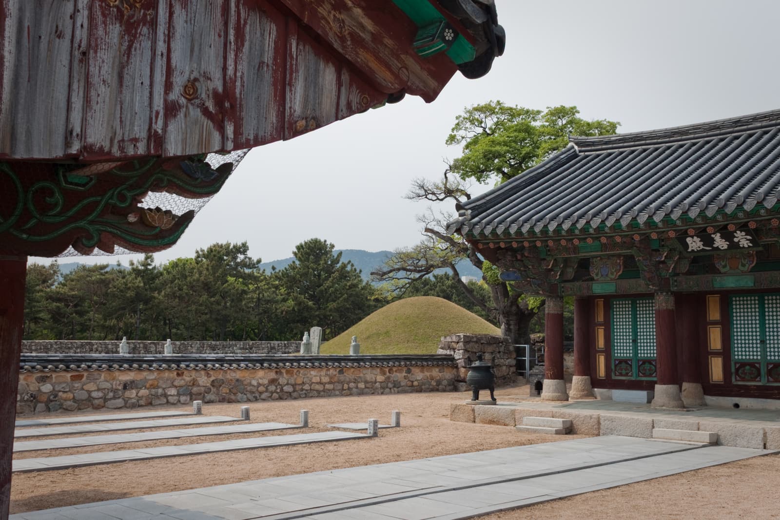 A traditional Korean temple with ornate, colorful wooden eaves and tiled roofs. The temple grounds have paved pathways leading to the ancient structures, and a royal burial mound can be seen in the background, highlighting the sacred and historical nature of the site.