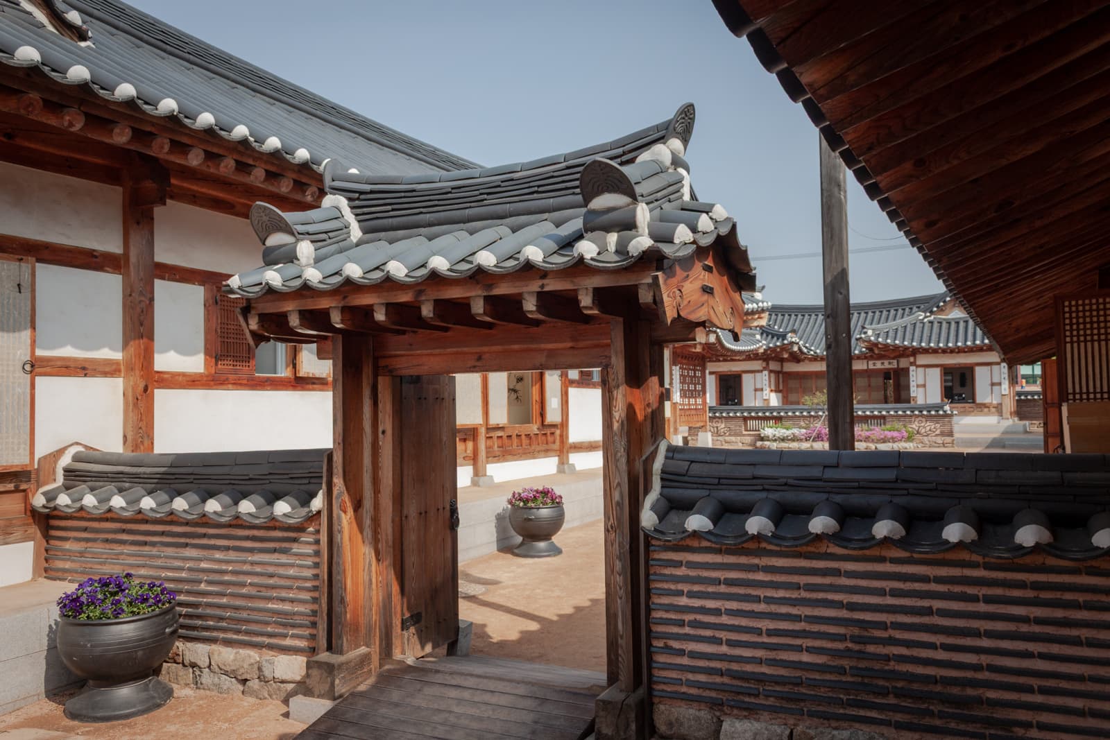 A traditional Korean courtyard with wooden structures and stone-tiled roofs, capturing the serene architecture of a hanok (Korean traditional house). The wooden gate leads into the courtyard, showcasing the harmonious blend of wood and stone elements typical of historic Korean homes.