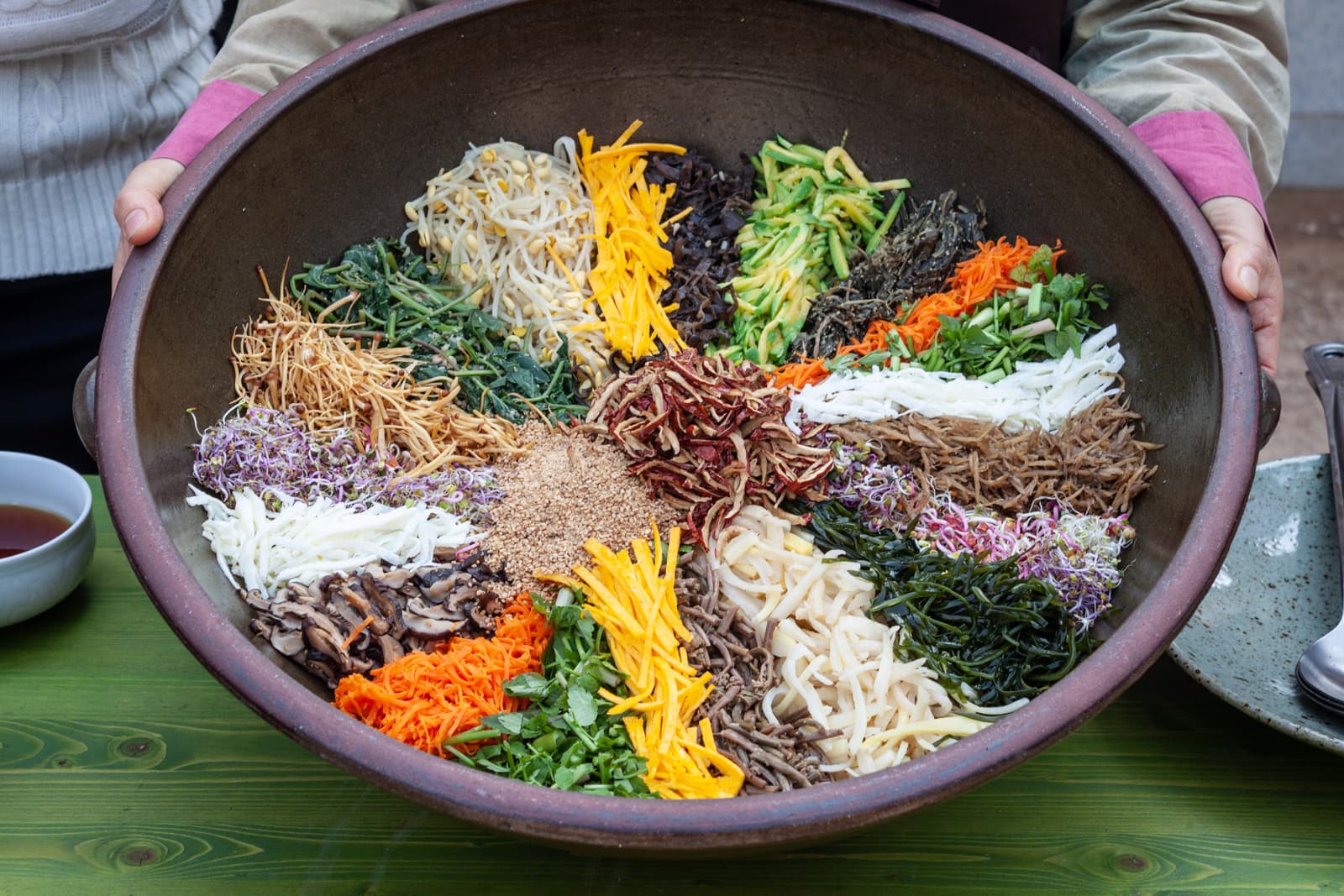 A large bowl filled with an assortment of vibrant vegetables arranged in sections. The colorful ingredients include shredded carrots, mushrooms, sprouts, and greens, forming a visually striking display, likely for a traditional dish like bibimbap or a large vegetable salad.