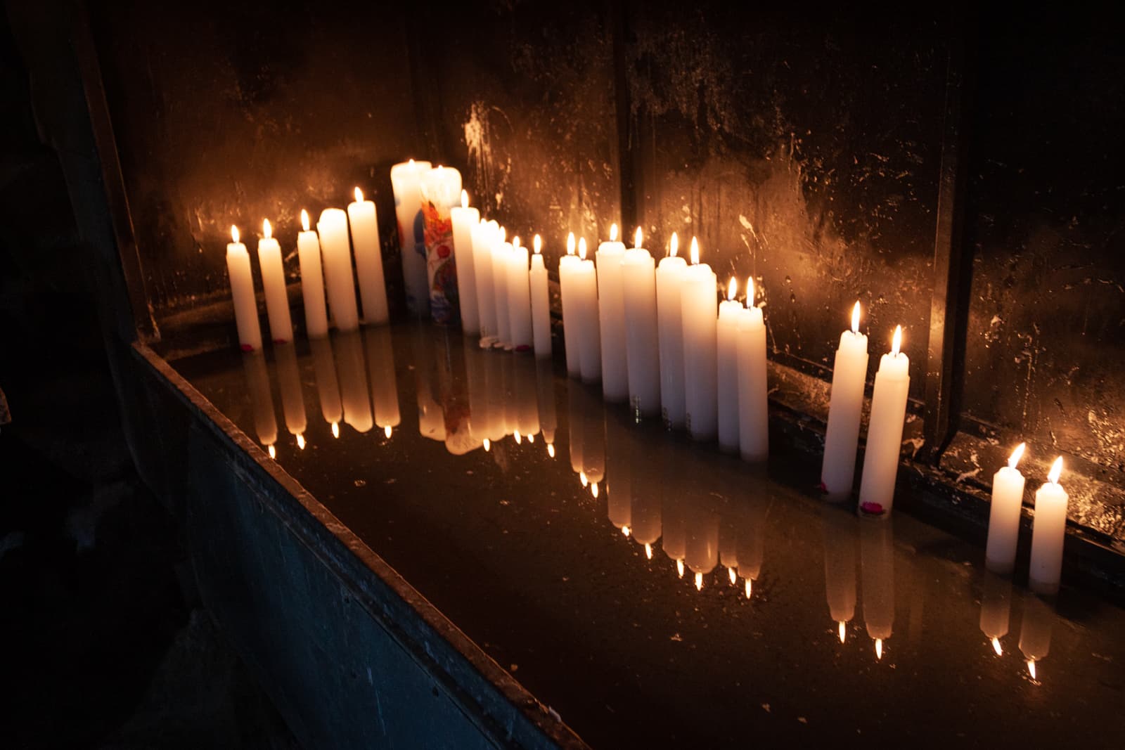 A dimly lit space featuring rows of white candles burning brightly, their reflection visible in the water below, creating a peaceful and reverent atmosphere.