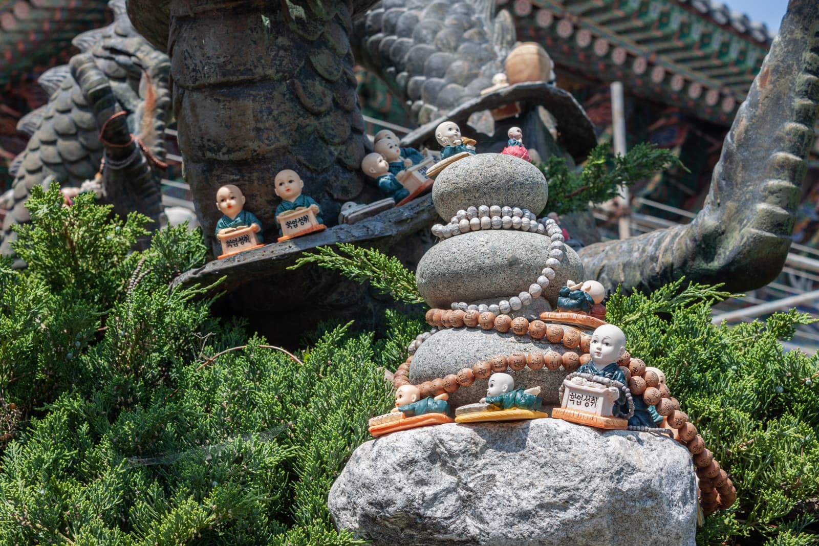 Small figurines of monk children placed on a stone with prayer beads draped over it, surrounded by lush green bushes and a dragon statue in the background, part of a serene temple setting.