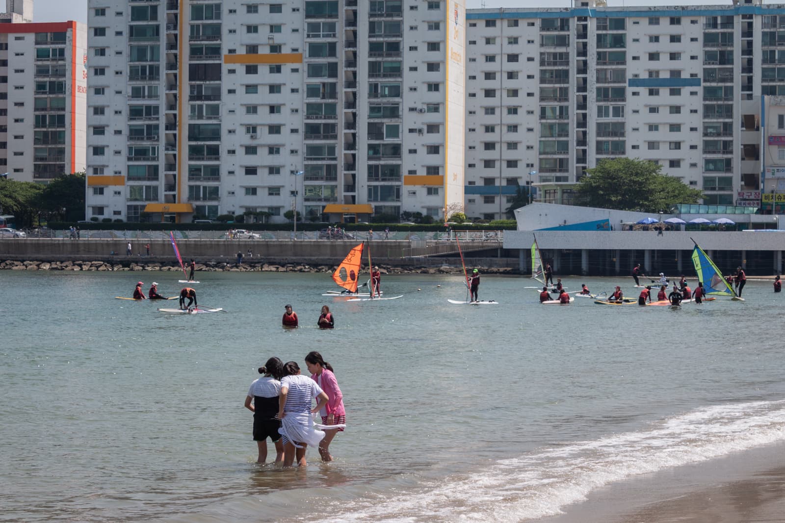 A beach scene with people wading in the shallow water and groups of individuals learning to windsurf or paddleboard. In the background, large apartment buildings line the beachfront, contributing to a blend of urban and coastal life.