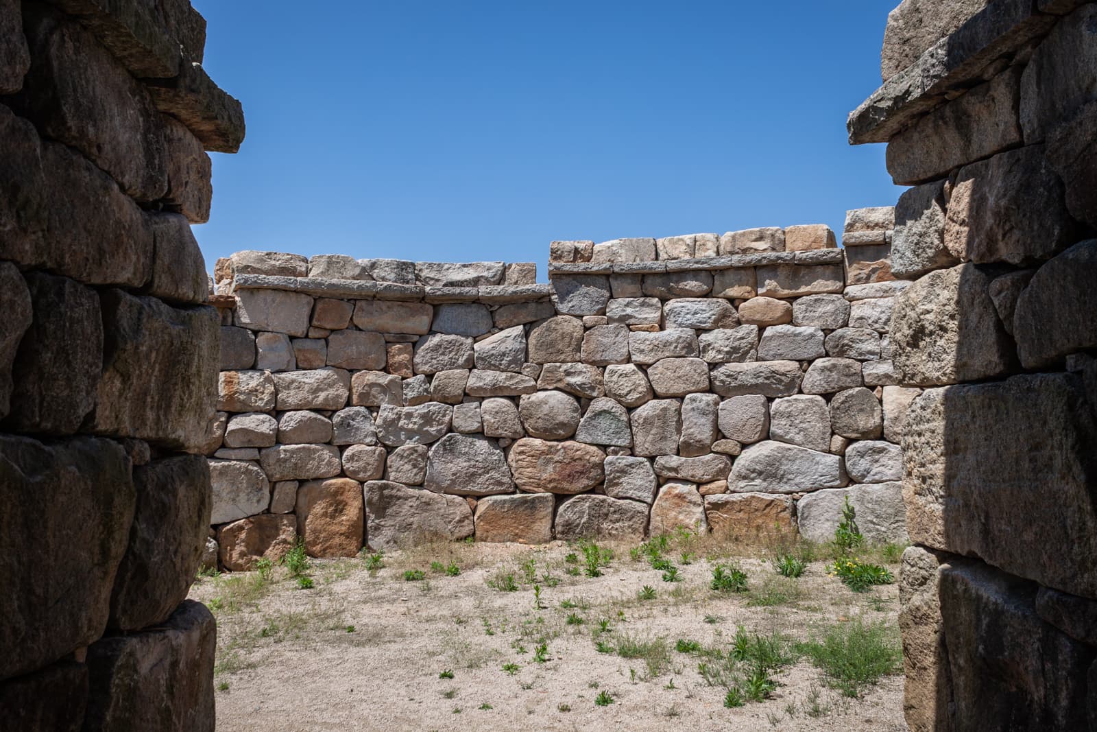 A view of an ancient stone enclosure with thick, weathered stone walls under a clear blue sky. The interior ground is sandy with sparse patches of grass and small plants. The rough, irregular stones are tightly fitted together, forming a protective boundary that suggests this is part of a historical fortification or structure.