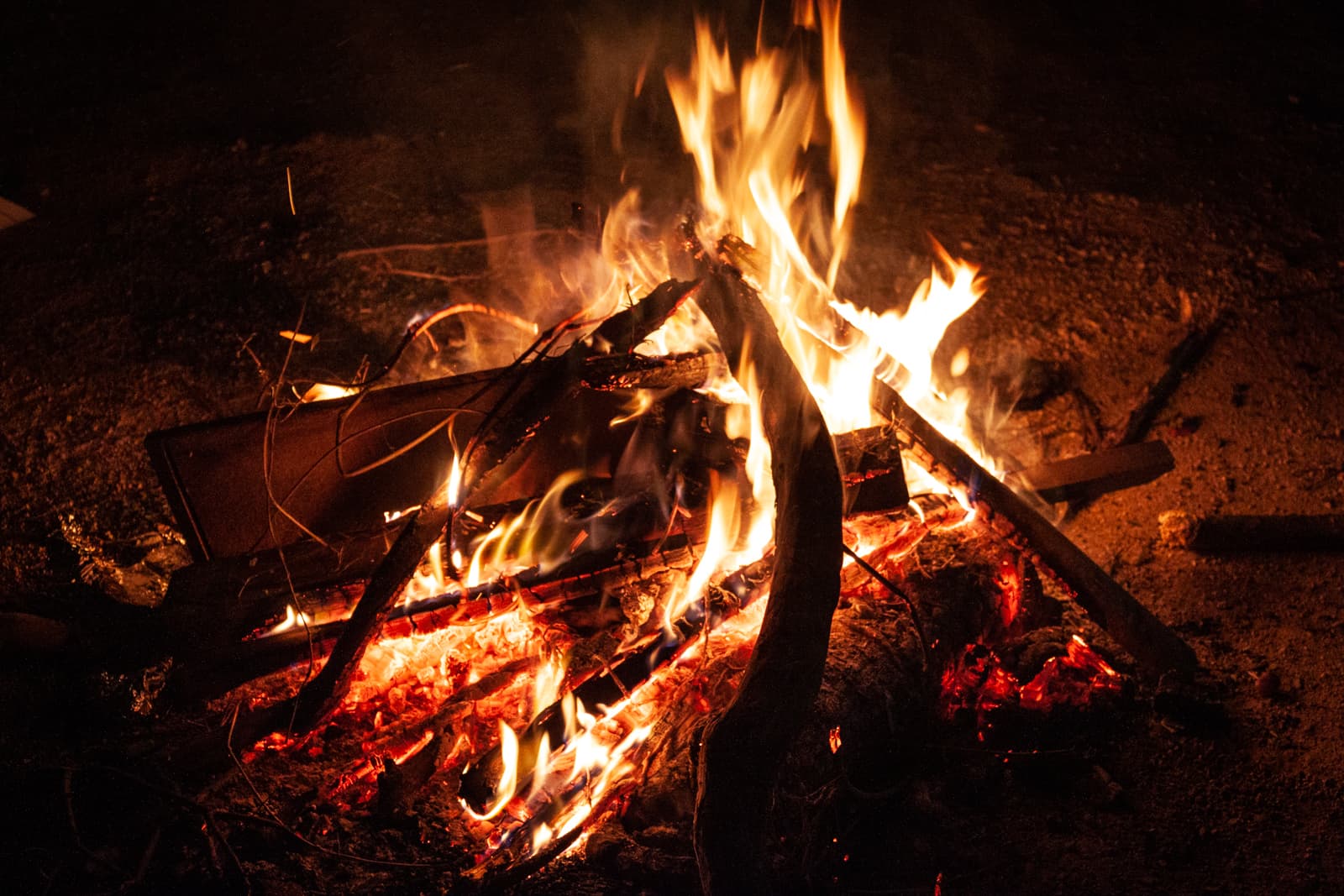 A campfire burning brightly at night, with logs and sticks crackling in the flames. The firelight illuminates the surrounding ground, creating a warm and rustic scene.