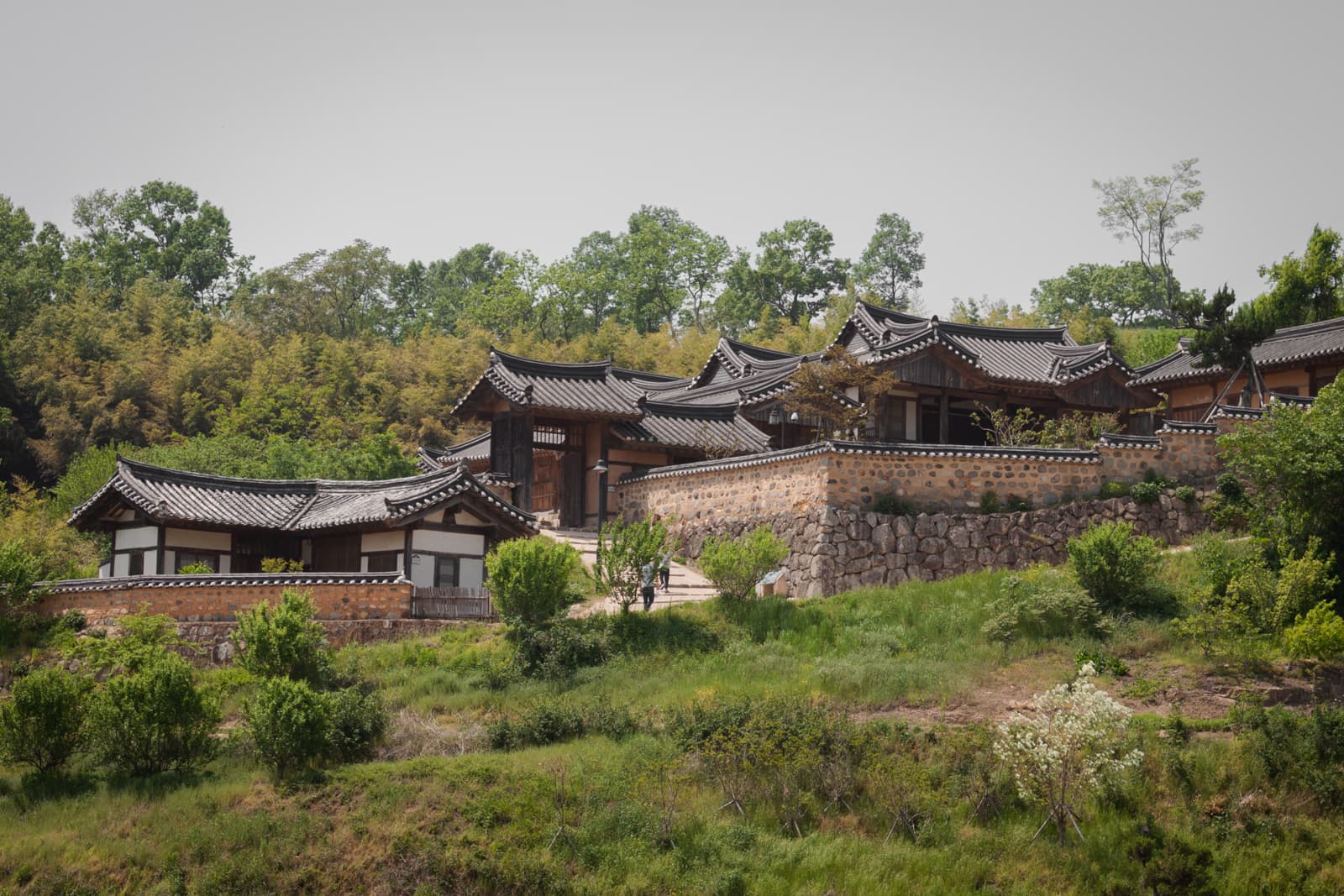 A hillside village of traditional Korean hanok homes, with stone walls and tiled roofs, surrounded by greenery. The homes are nestled into the landscape, evoking a serene and historical ambiance.