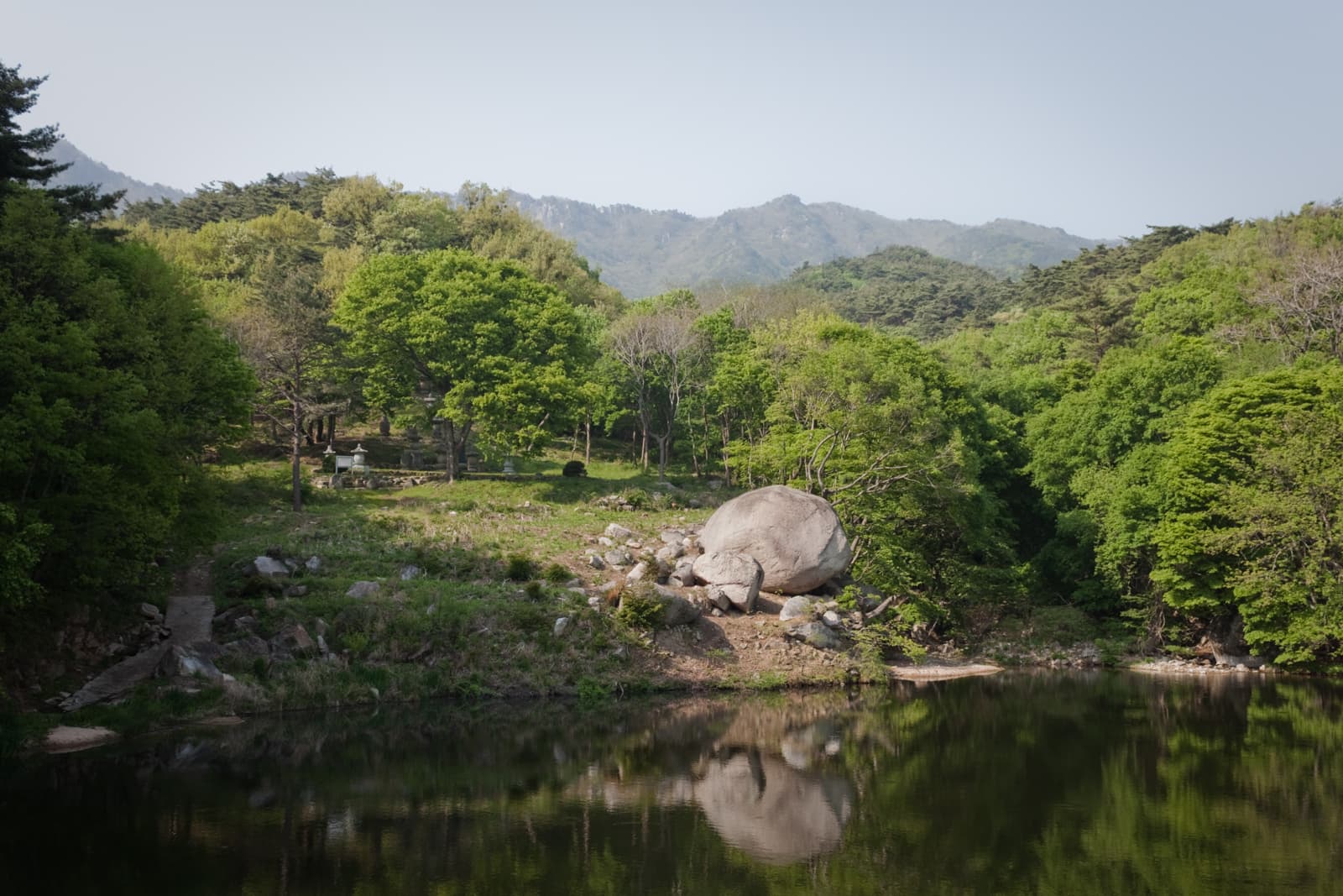 A tranquil scene of a reflective pond surrounded by lush green trees, with mountains in the background. A large boulder sits near the edge of the water, enhancing the peaceful, natural setting.