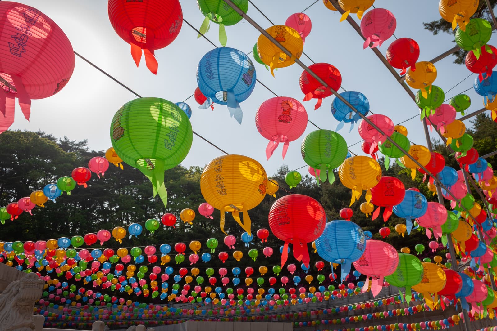Rows of colorful lanterns in shades of red, green, yellow, and blue are strung up against the sky. The festive lanterns hang in a canopy over a temple courtyard, adding a lively and vibrant atmosphere.