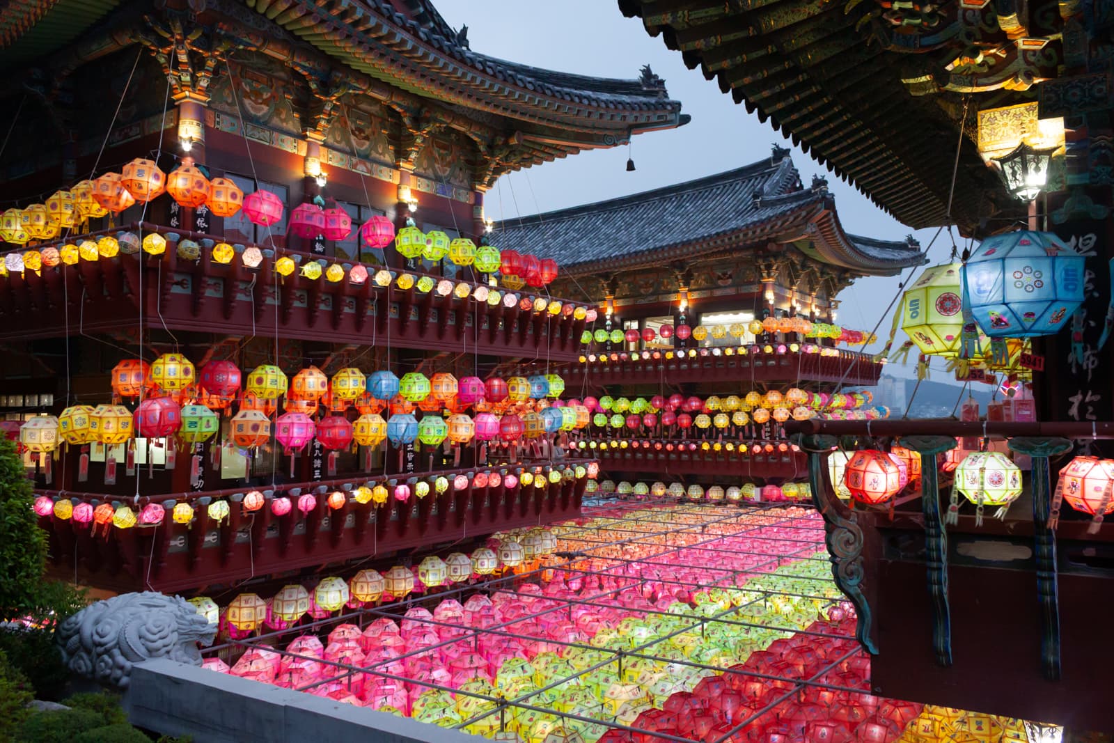 A vibrant display of colorful lanterns in various shades of pink, yellow, green, and orange, strung up around a traditional Korean temple building, giving the scene a festive and illuminated appearance as dusk settles in.