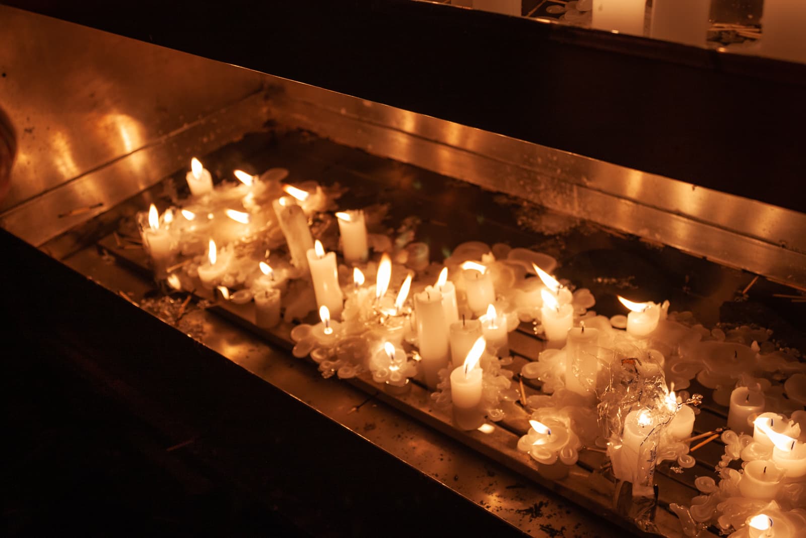 A row of candles melting and burning softly in a metal tray, their warm light reflecting in the melted wax, creating a serene atmosphere.
