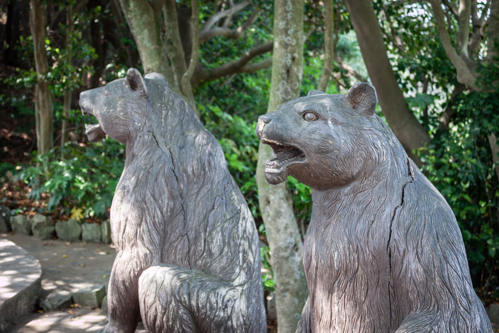 Two wooden bear sculptures with their mouths open, standing side by side in a wooded area.