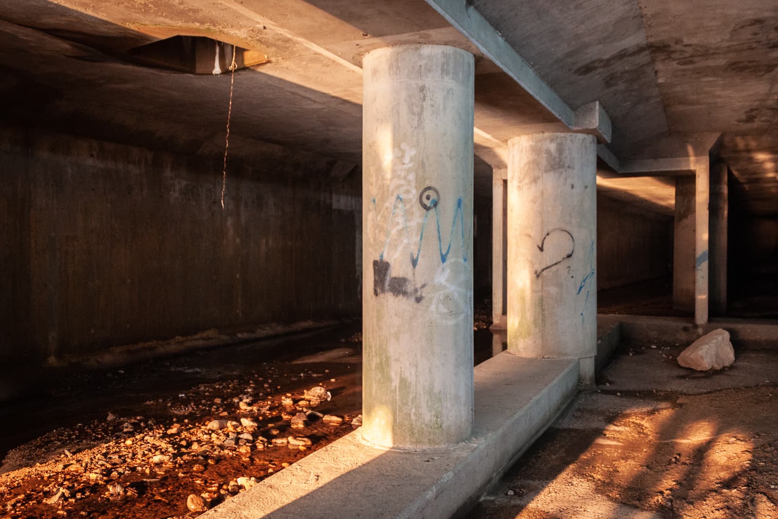 The interior of a concrete tunnel with large support pillars, some of which are covered in graffiti. The ground is rocky, with a small stream running through, illuminated by a warm glow from the light.