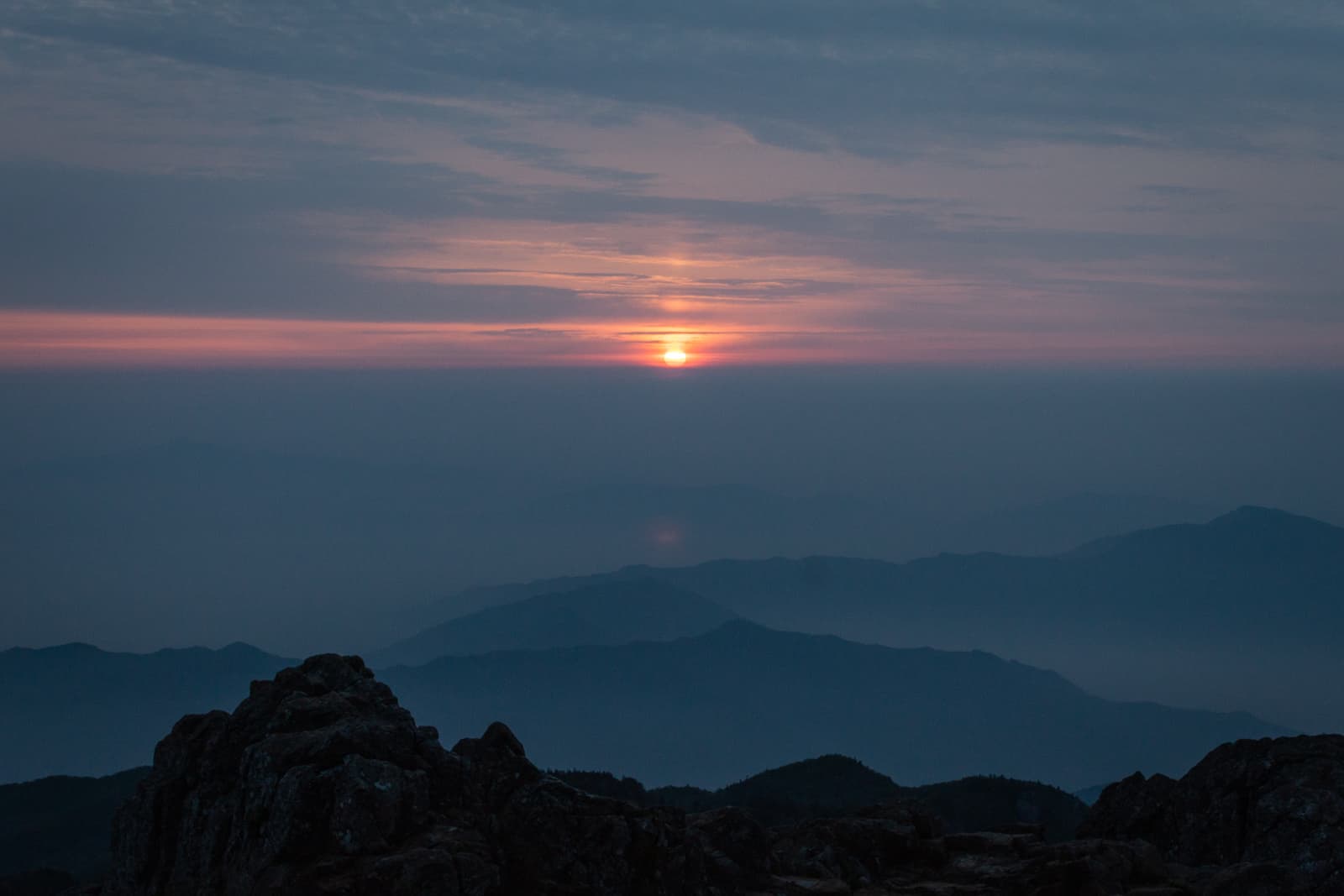 A colorful sunset casting soft light over a distant mountain range, with dark rocky silhouettes in the foreground.