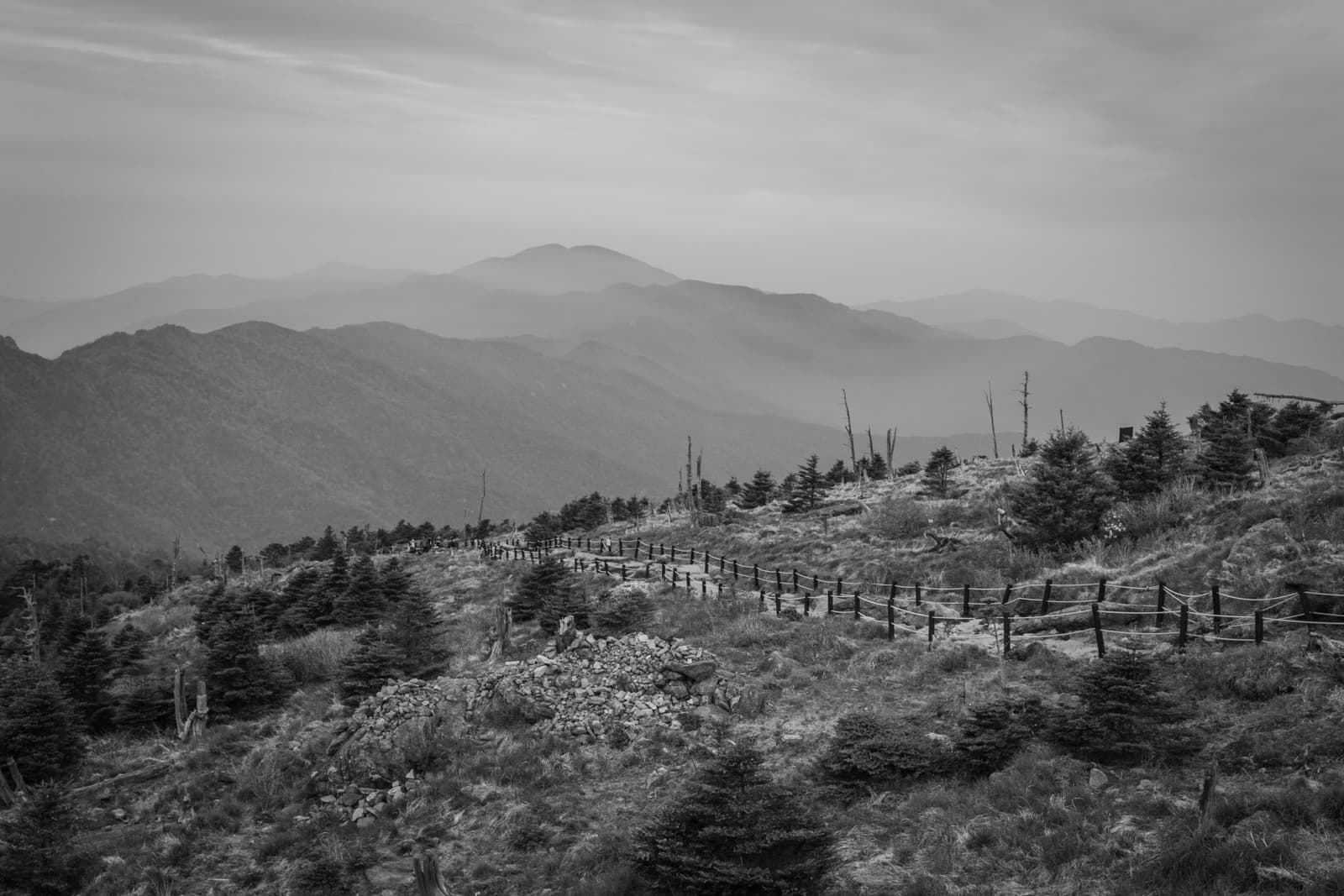 A black and white landscape of rolling mountains in the distance with a fence-lined path winding through a rocky hillside in the foreground.