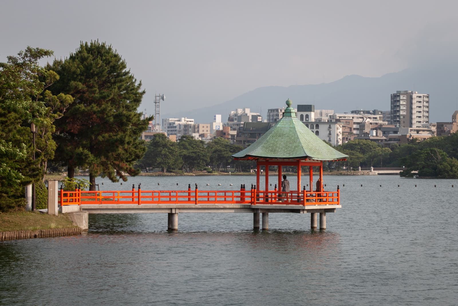 A traditional red gazebo with a green roof, built on a pier extending over a calm lake, with a city skyline and mountains visible in the distance.