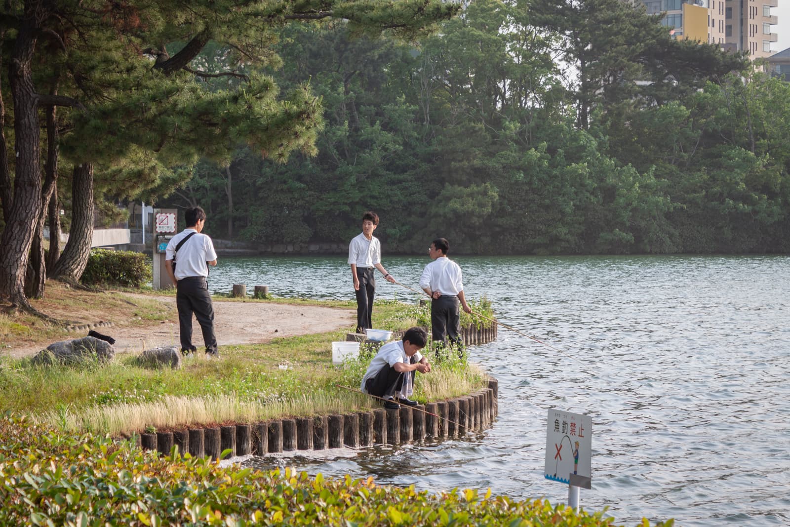 A group of young students, dressed in uniforms, standing and sitting by a lake, fishing with long rods near a shoreline lined with trees.