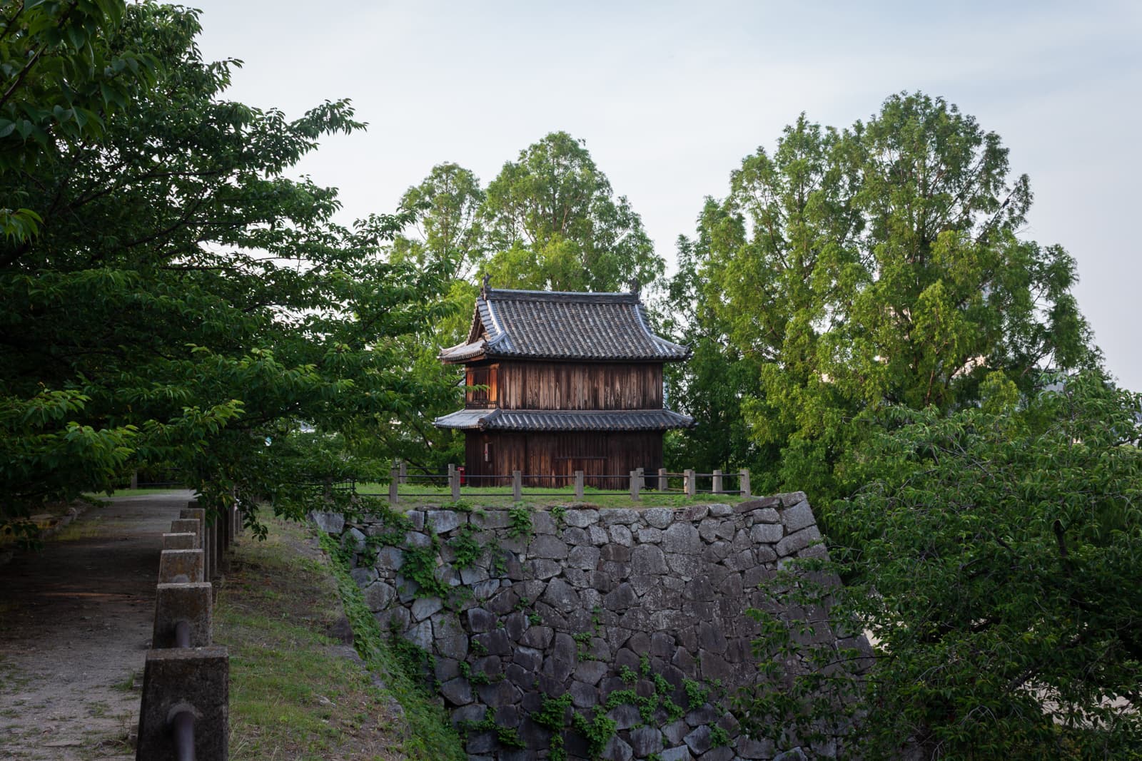 A traditional wooden structure with a tiled roof surrounded by lush green trees, sitting atop a stone wall.