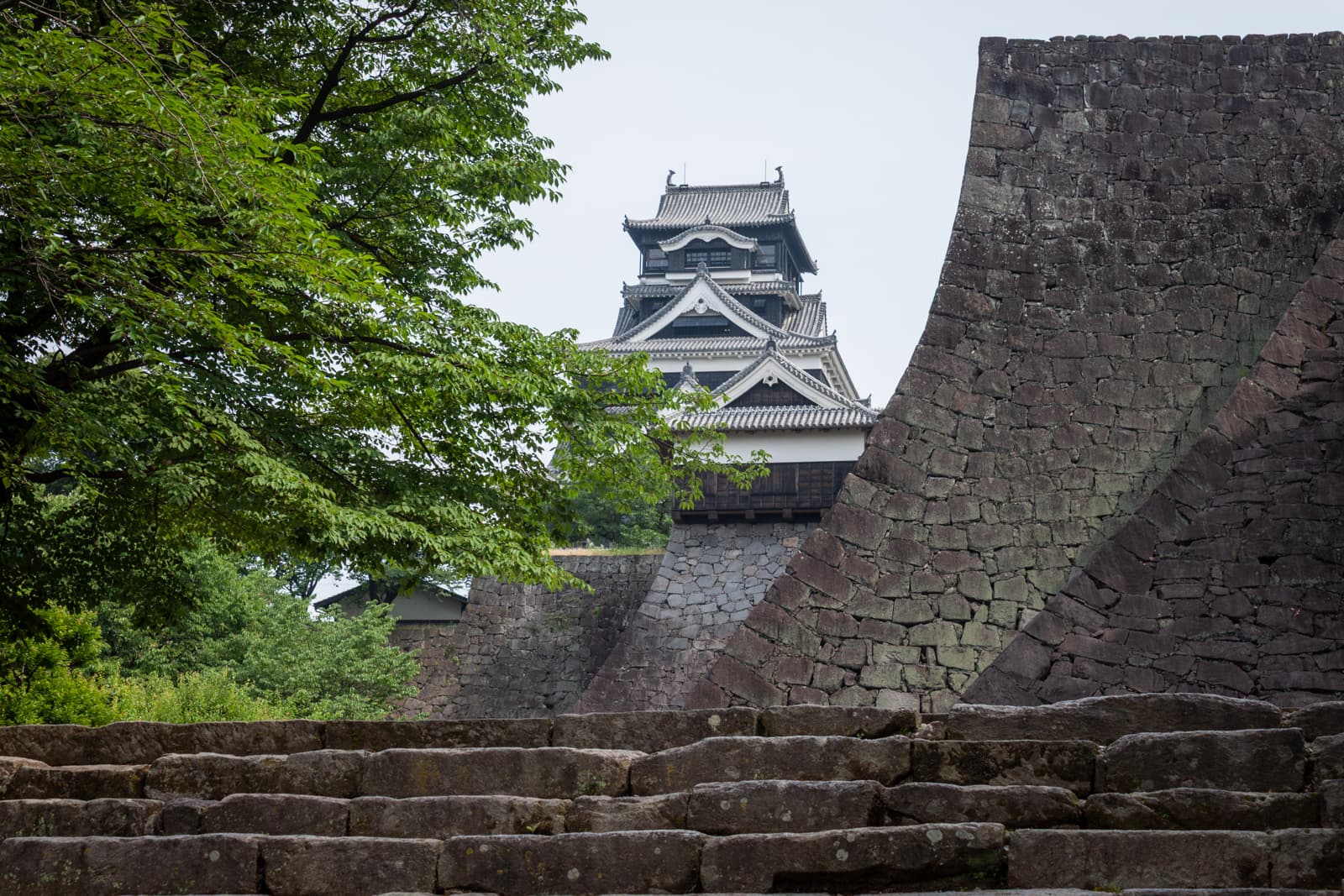 View of a traditional Japanese castle tower behind a steep stone wall, with steps leading up through a lush green landscape.