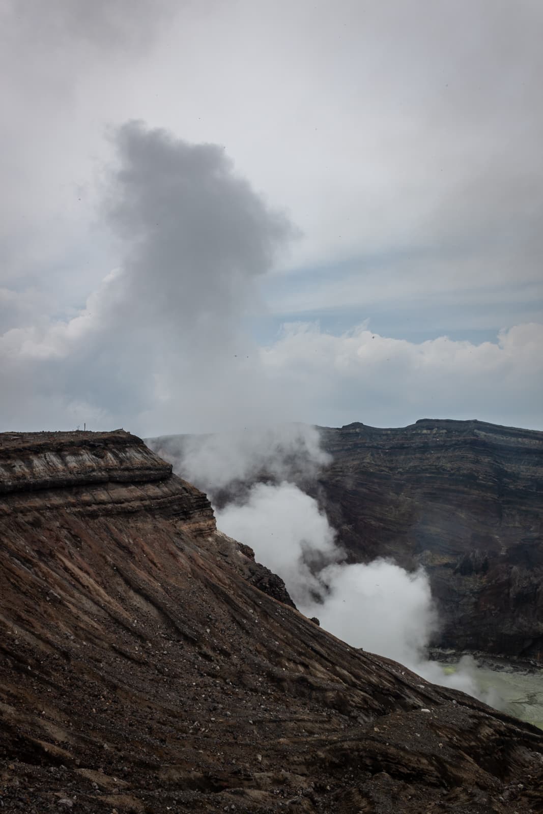 Clouds of steam emerge from the steep ridges of a volcanic landscape, against a cloudy sky.