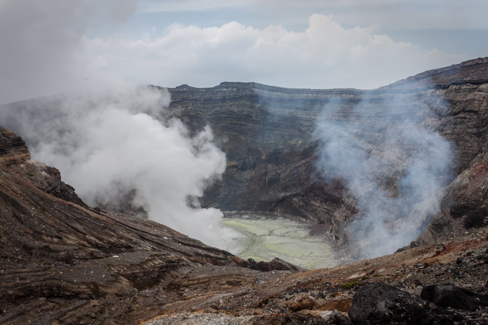 Thick clouds of steam rise from a rocky volcanic crater, with swirling vapors obscuring the crater floor filled with pale yellow water.
