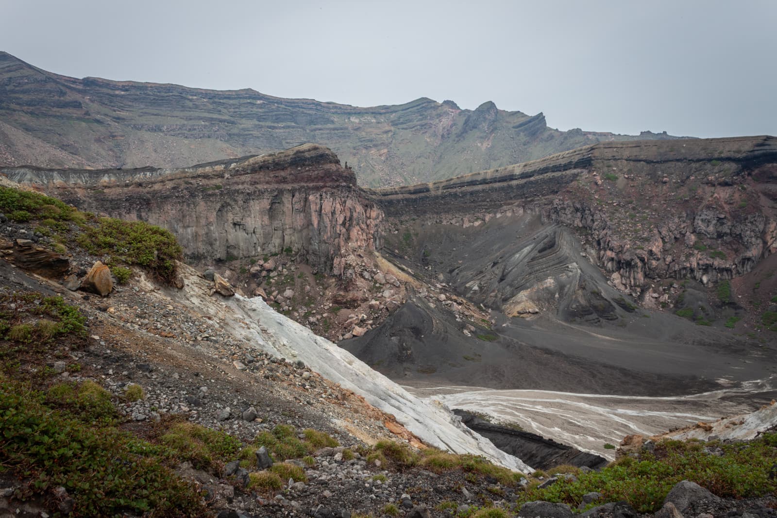 A view of rugged, steep cliffs and ash-laden slopes of a volcanic crater, showcasing layers of rock formations and patches of greenery.