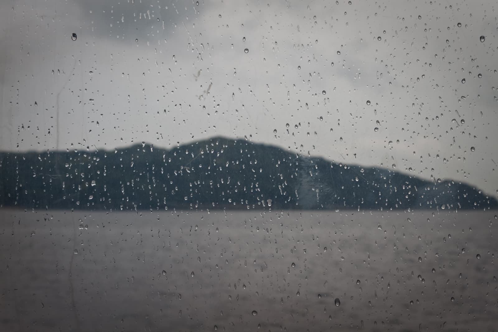 Raindrops cover a window, blurring the view of a distant island across a body of water, under a cloudy, overcast sky.