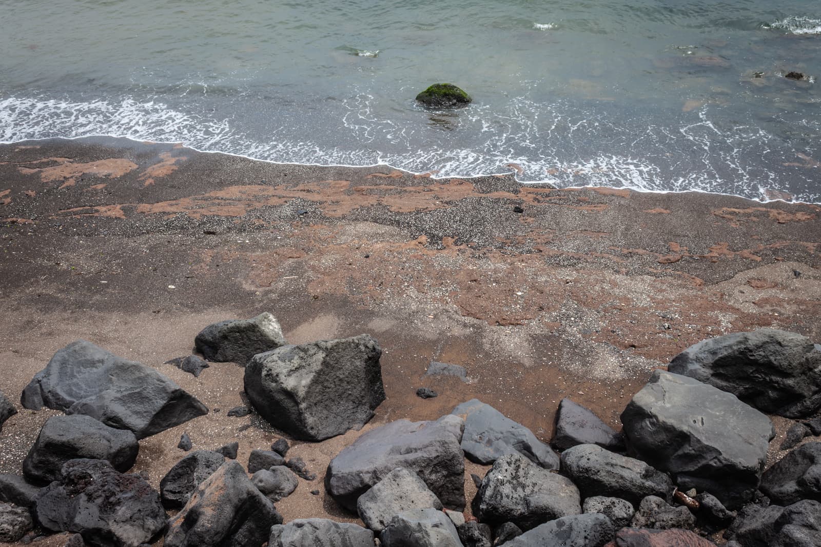 A shoreline with black sand and large dark rocks scattered along the water’s edge, with gentle waves lapping the shore.