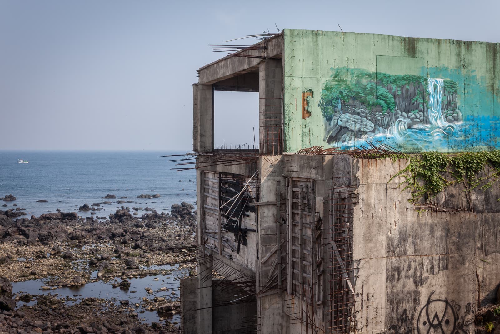 A partially crumbling building with a colorful mural of a waterfall, standing on the rocky shore of the sea.