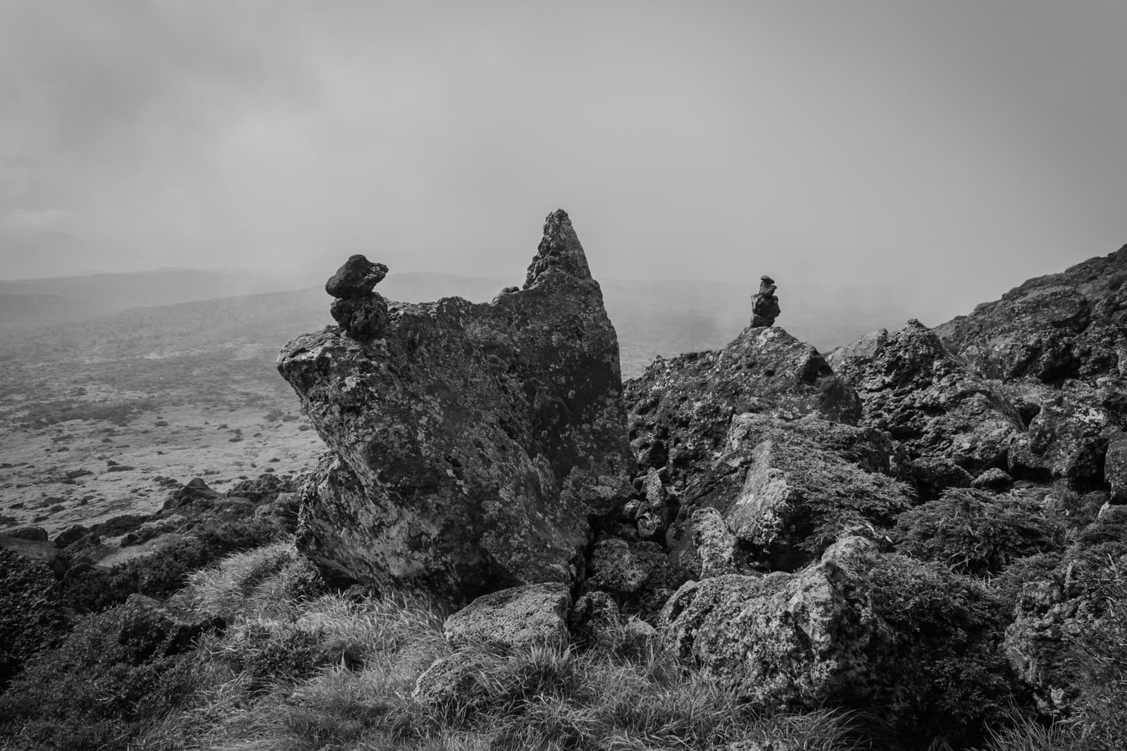 Two large, weathered rock formations stand atop a grassy hill, shrouded in mist. The rocks are covered with moss, and the hazy horizon gives the image a mysterious, otherworldly tone.