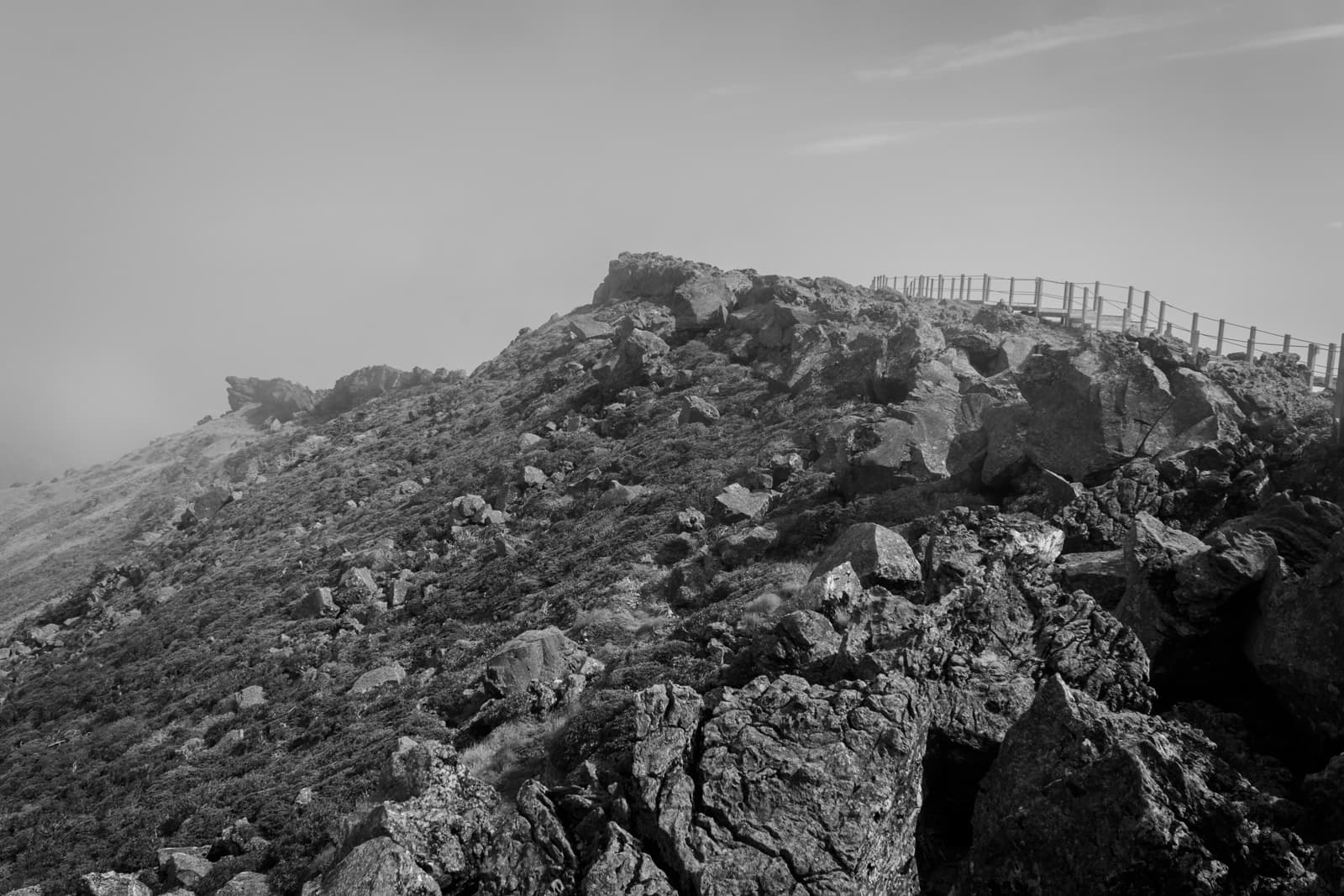 A rugged hillside covered with large boulders and low vegetation, with a faint pathway and fencing along the crest of the hill. The black-and-white image captures a serene yet harsh atmosphere.