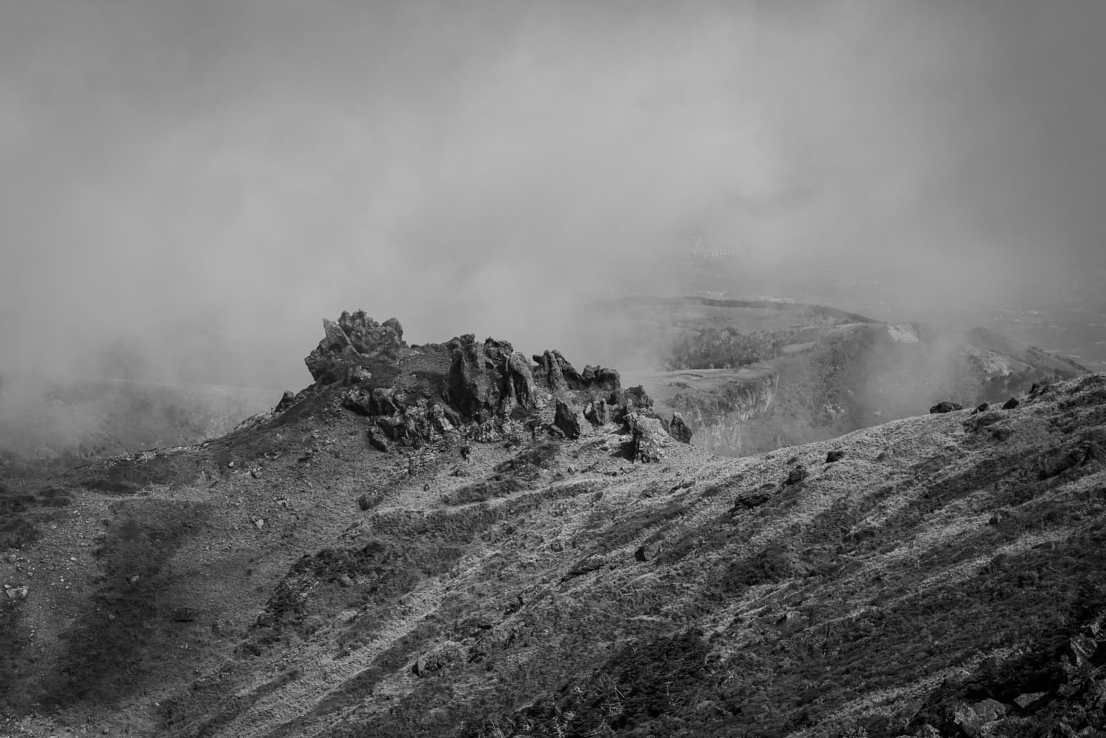 A rocky ridge with jagged outcrops, partially obscured by thick fog. The terrain is barren, with sharp rock formations rising against a cloudy sky in this black-and-white landscape.